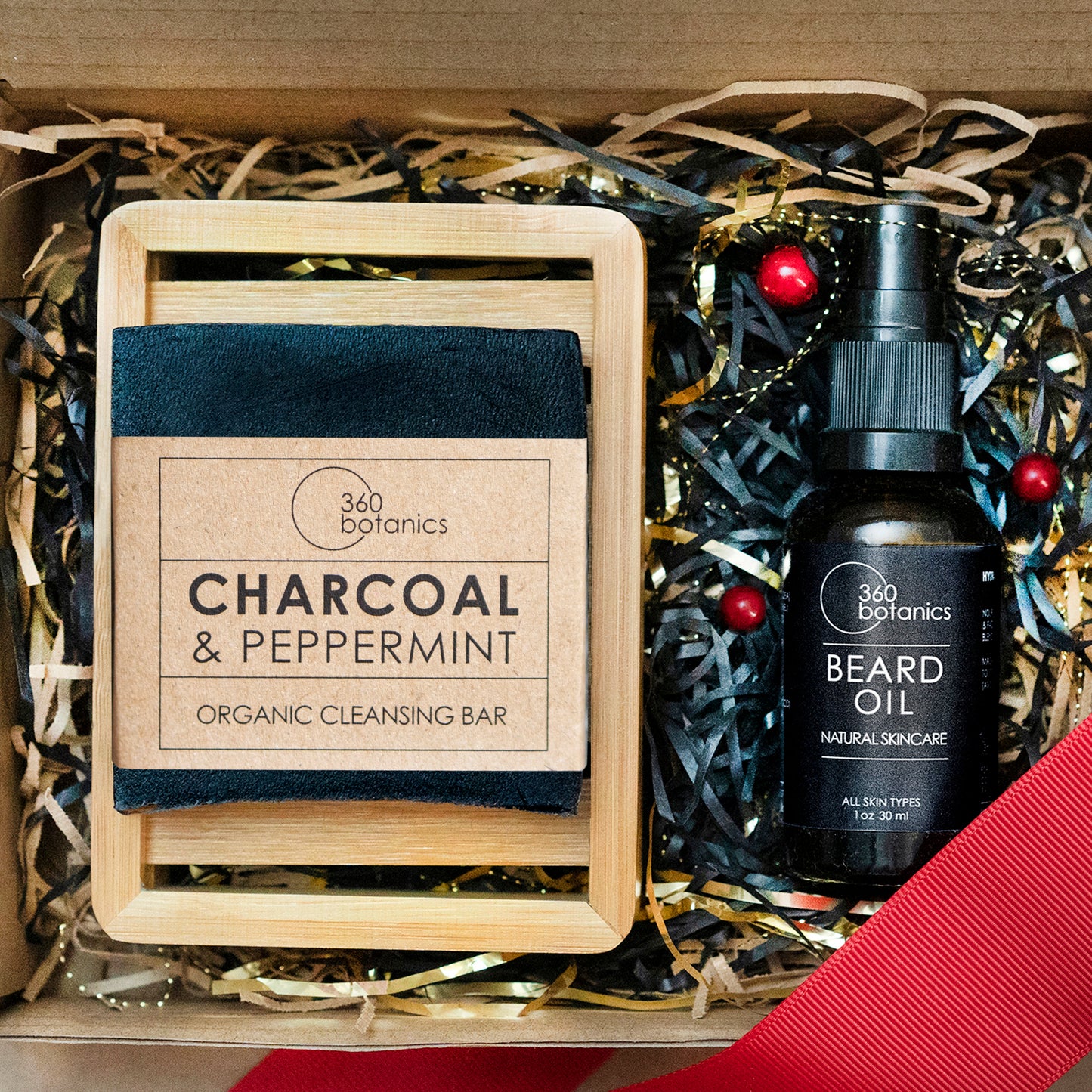 Festive 360 Botanics gift set featuring a Charcoal & Peppermint Organic Cleansing Bar on a bamboo tray, next to a bottle of Beard Oil. The items are nestled in a bed of black and gold crinkle paper with red berry accents, exuding a luxurious and holiday-ready appeal