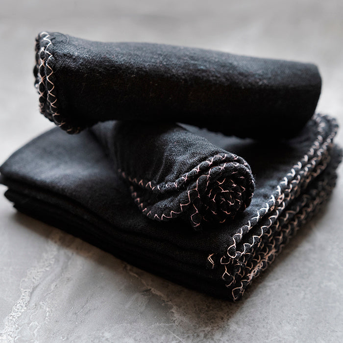  A rolled and a folded stack of black muslin cloths with decorative edge stitching, displayed on a marbled grey surface.