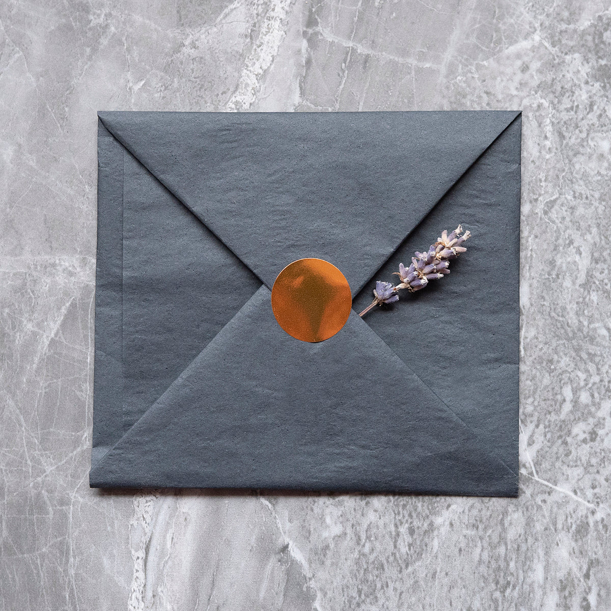 A dark gray envelope sealed with a copper-colored sticker and decorated with a sprig of lavender, placed on a marble surface, conveying an elegant and serene correspondence theme.