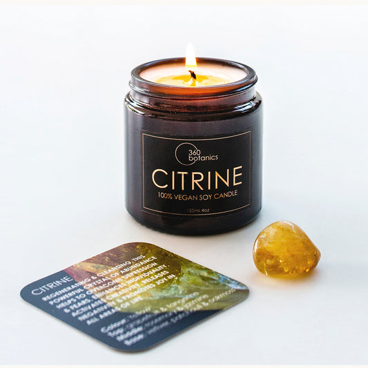 A lit 360 Botanics Citrine 100% vegan soy candle in a dark glass jar, next to a polished citrine stone and an information card about citrine properties on a white surface.