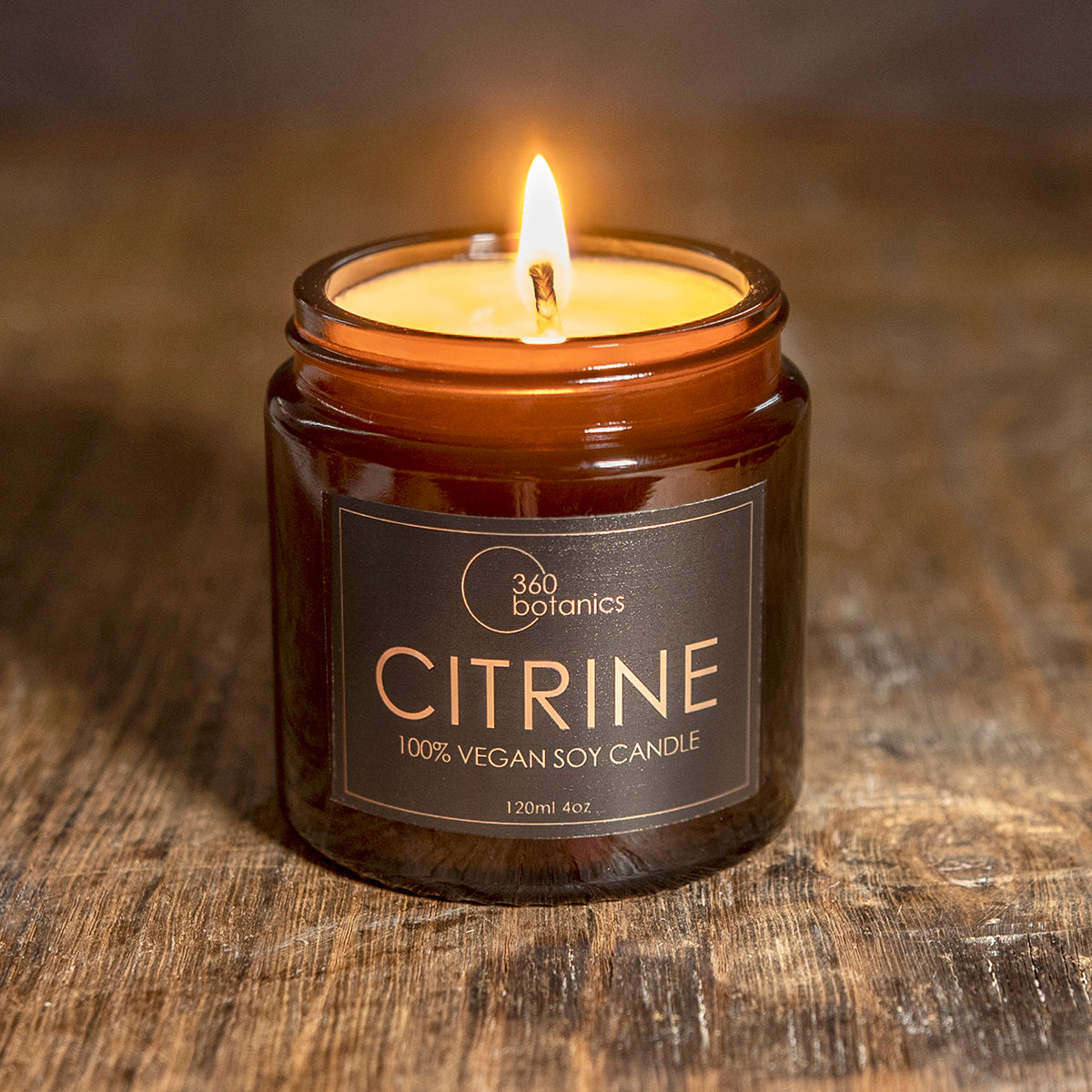 A lit 360 Botanics Citrine 100% vegan soy candle in a brown glass jar, with a glowing flame, placed on a rustic wooden surface.