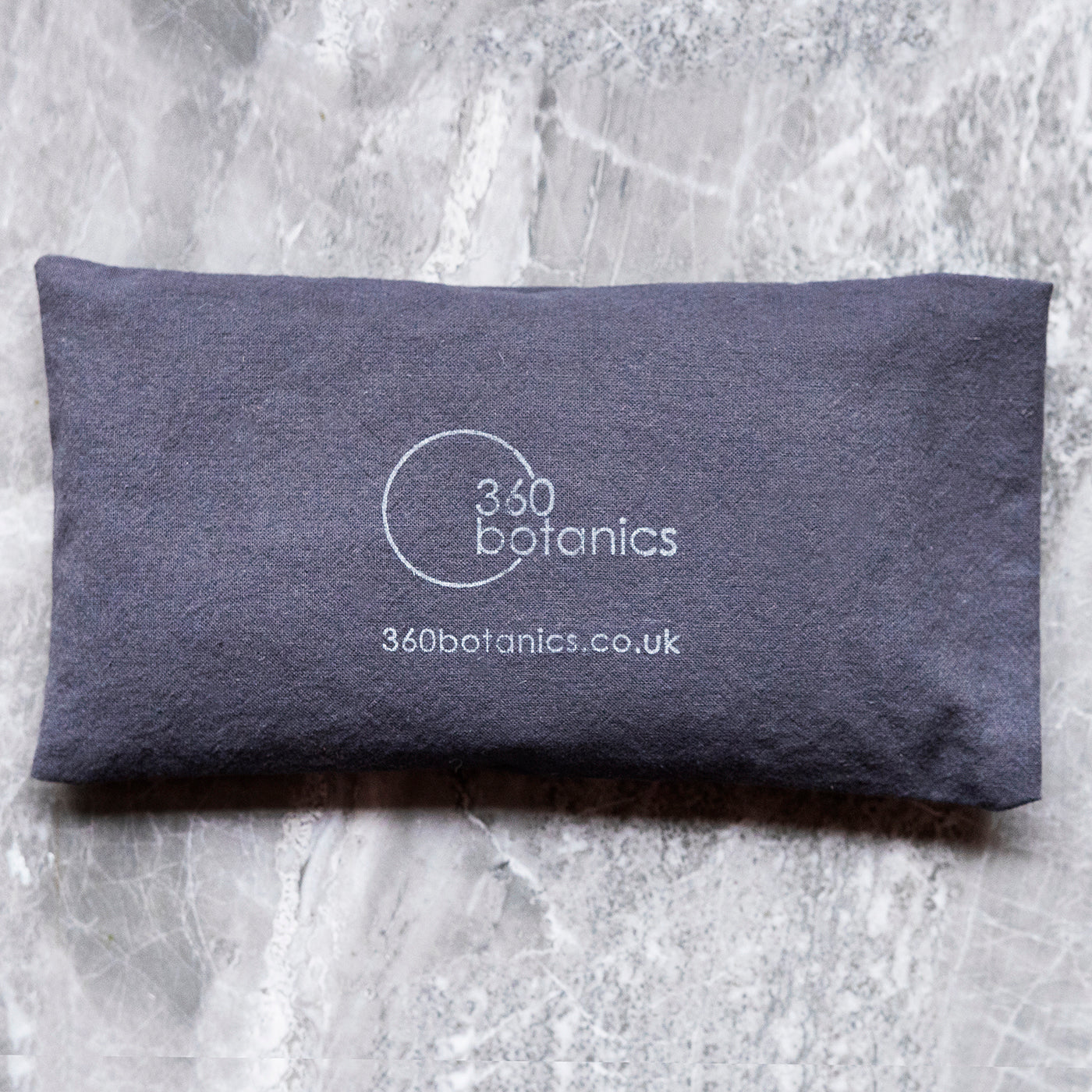  A navy blue eye pillow with the white logo "360 Botanics" and the website "360botanics.co.uk" on a marbled grey background.