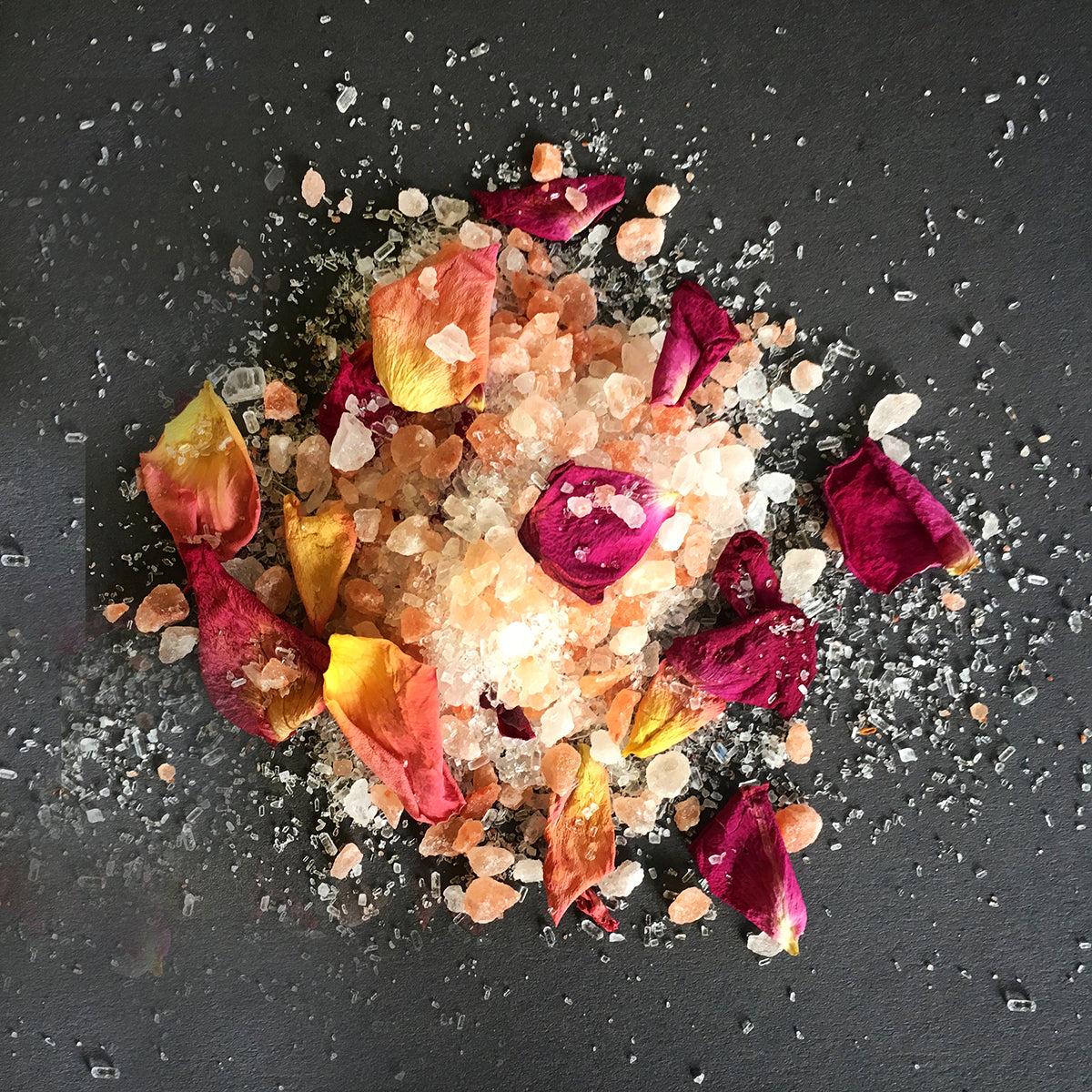 A close-up view of a luxurious mixture of bath salts and dried flower petals arranged on a dark, textured surface. The salt crystals vary in color from clear to peach and amber tones, while the flower petals are rich in color, including shades of deep burgundy, bright yellow, and warm orange. The scattered arrangement gives an impression of a relaxing and aromatic bath experience