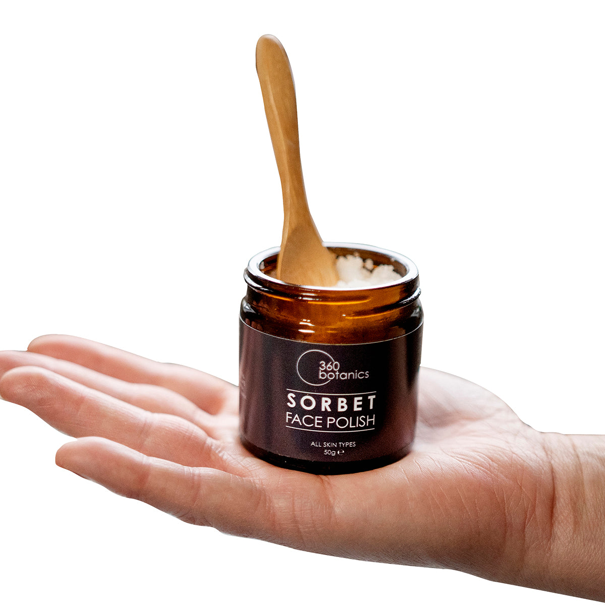 Sorbet face polish in amber jar on palm of hand, woodedn spoon coming out of jar, white backgroun