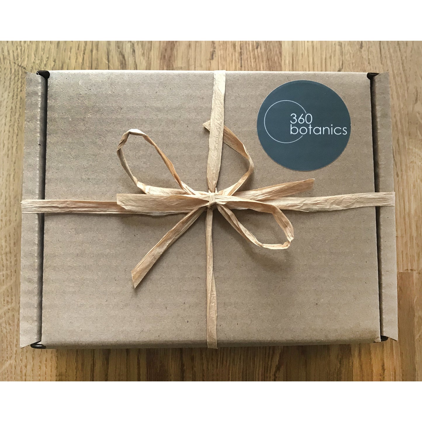 Eco-friendly packaging from 360 Botanics featuring a brown kraft paper box tied with a natural raffia ribbon. The company's logo is prominently displayed on a circular, teal sticker on the top of the box, suggesting a thoughtful and sustainable product presentation