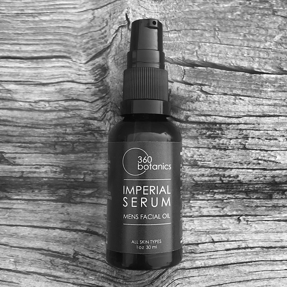 A bottle of 360 Botanics Imperial Serum men's facial oil on a rustic wooden background. The amber glass bottle has a black label and spray nozzle, and is presented in grayscale tones.