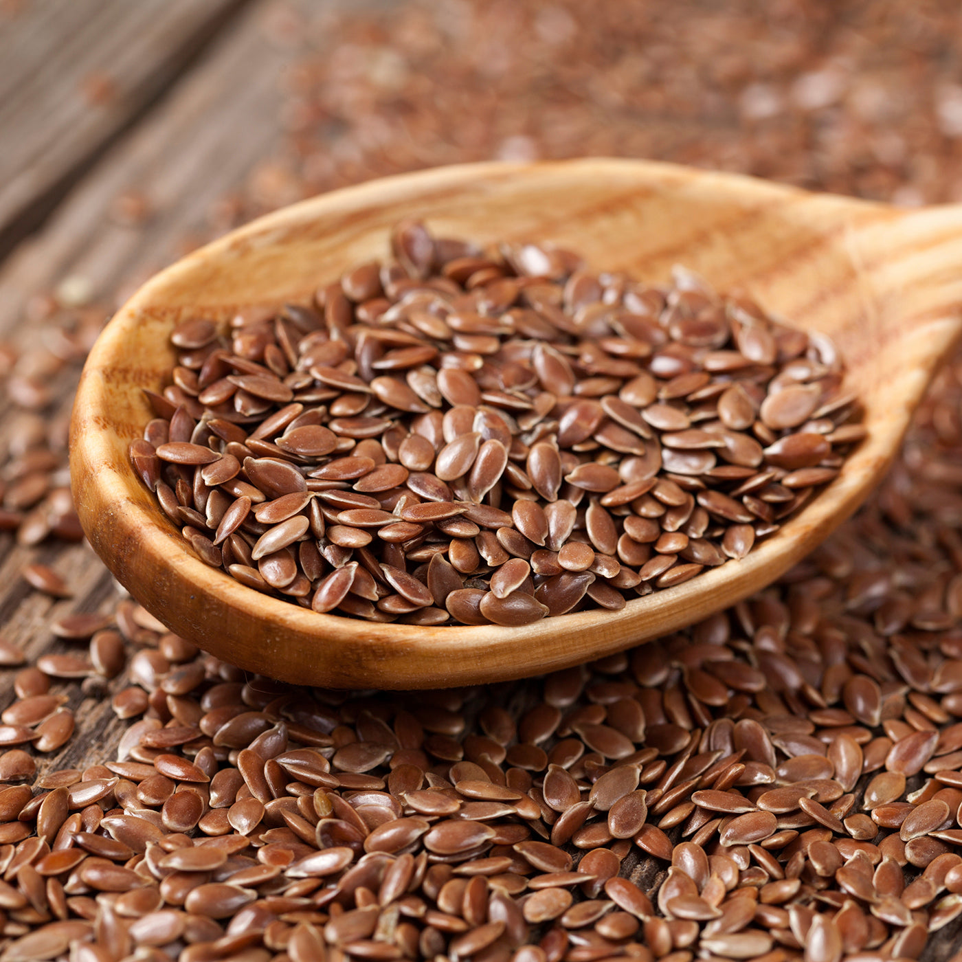  A wooden spoon overflowing with brown flaxseeds set on a backdrop of more flaxseeds on a rustic wooden surface.