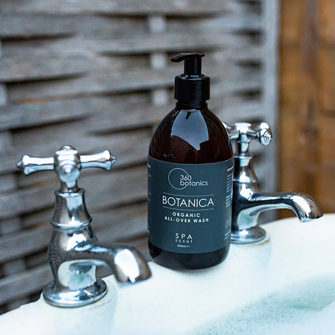 A bottle of 360 Botanics Botanica Organic All-Over Wash with a pump dispenser, labeled "SPA SCENT" in 500ml size, placed next to a vintage style sink with chrome taps, against a wooden plank backdrop.