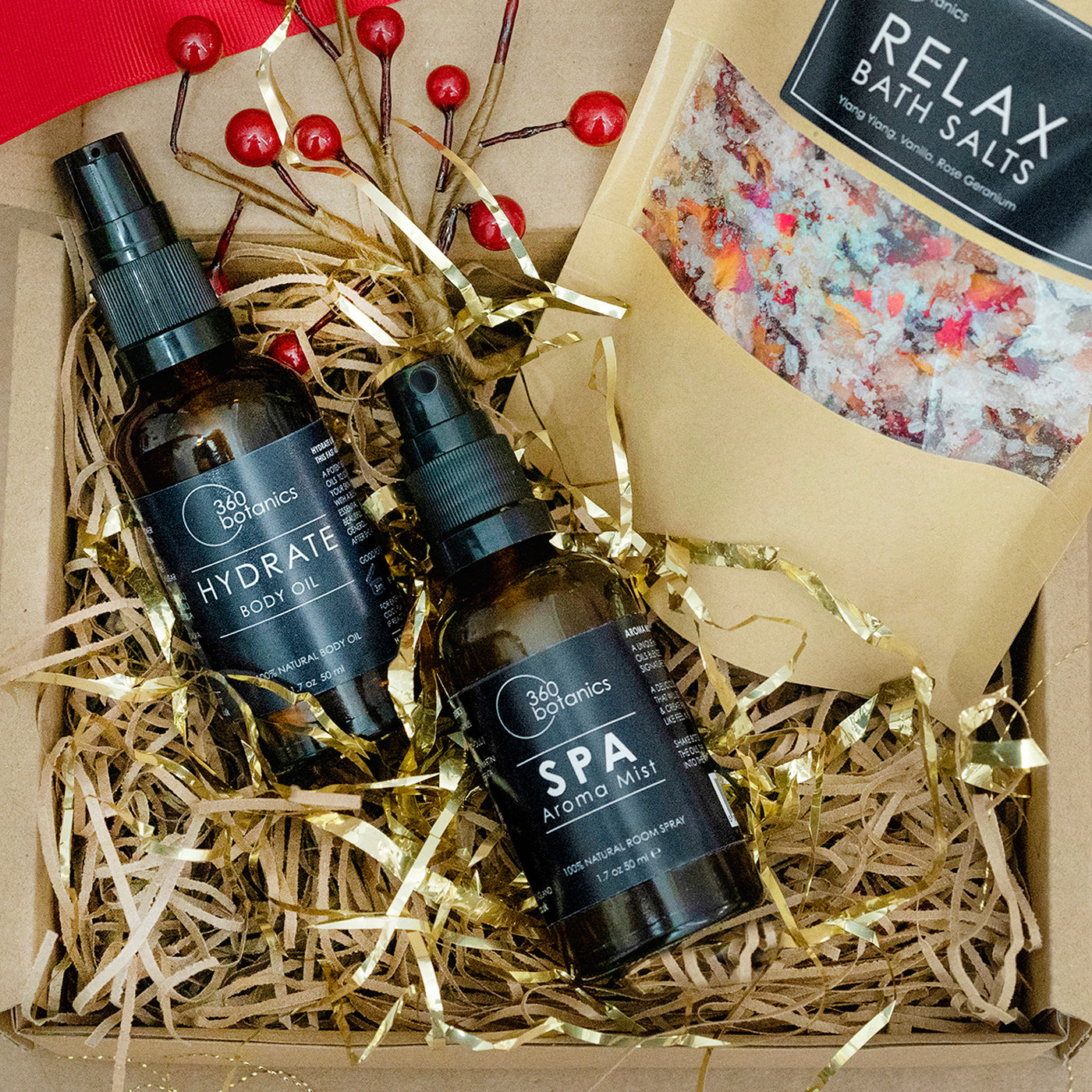 A gift box filled with shredded gold paper featuring two "360 Botanics" products: "HYDRATE Body Oil" and "SPA Aroma Mist," with a pouch of "RELAX Bath Salts" partially visible and decorative red berries on branches adding a festive touch