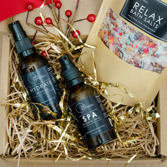 A gift box filled with shredded gold paper featuring two "360 Botanics" products: "HYDRATE Body Oil" and "SPA Aroma Mist," with a pouch of "RELAX Bath Salts" partially visible and decorative red berries on branches adding a festive touch