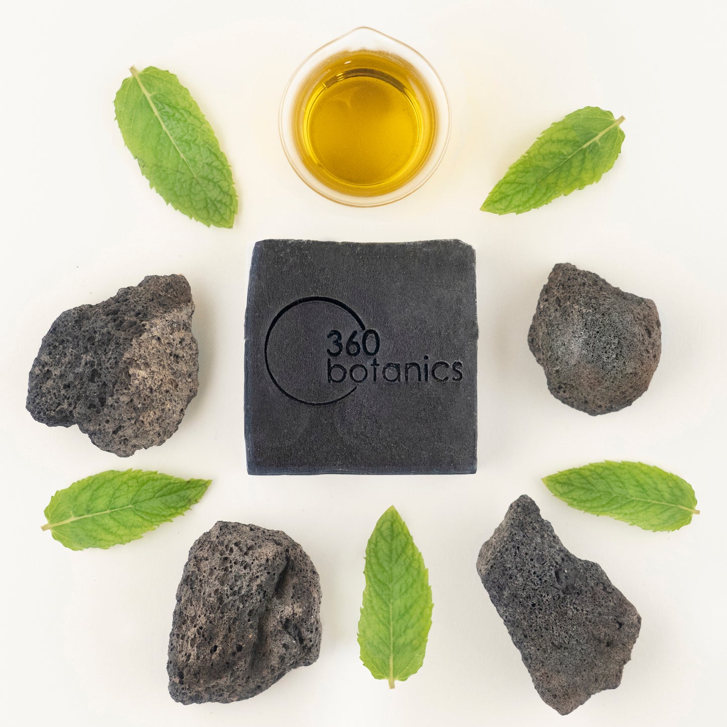 A top-down arrangement featuring a black 360 Botanics soap bar centered on a white background, surrounded by natural elements: fresh green leaves, volcanic rocks, and a small bowl of golden oil, creating a harmonious blend of textures and colors that emphasize the organic and earthy essence of the products