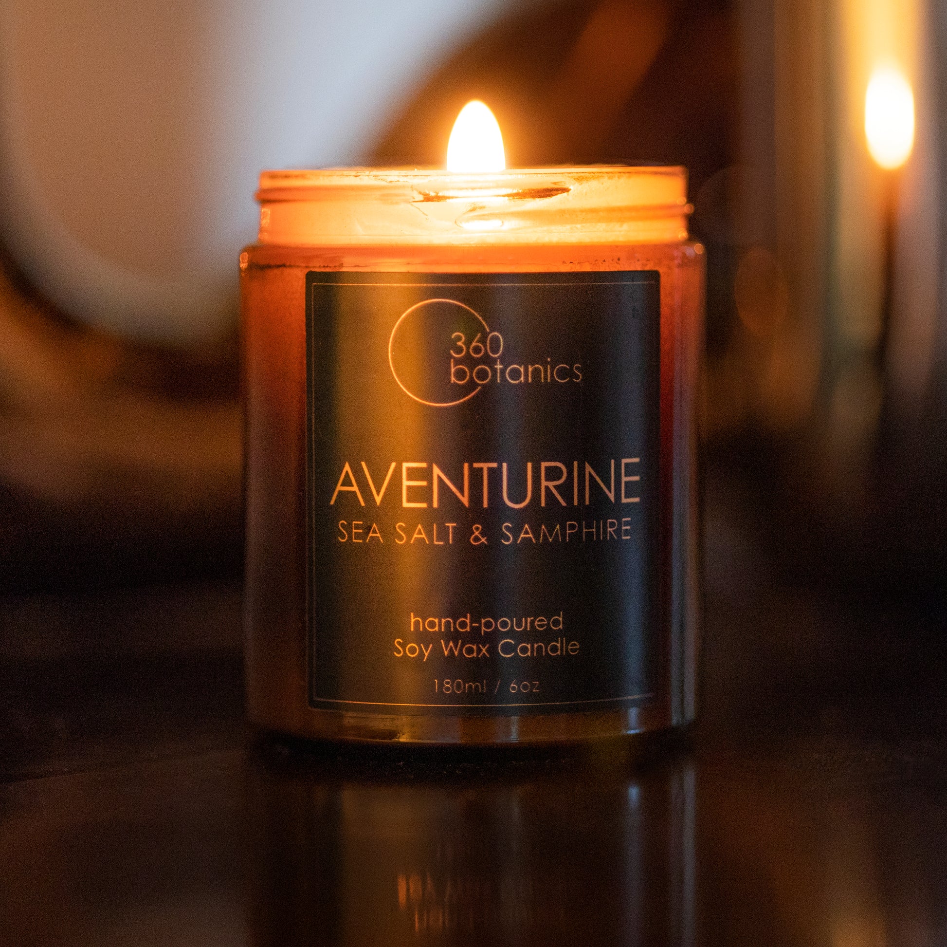 A glowing Aventurine hand-poured soy wax candle by 360 Botanics with 'Sea Salt & Samphire' scent, lit and casting a warm light in a dimly lit room, reflecting on a shiny surface