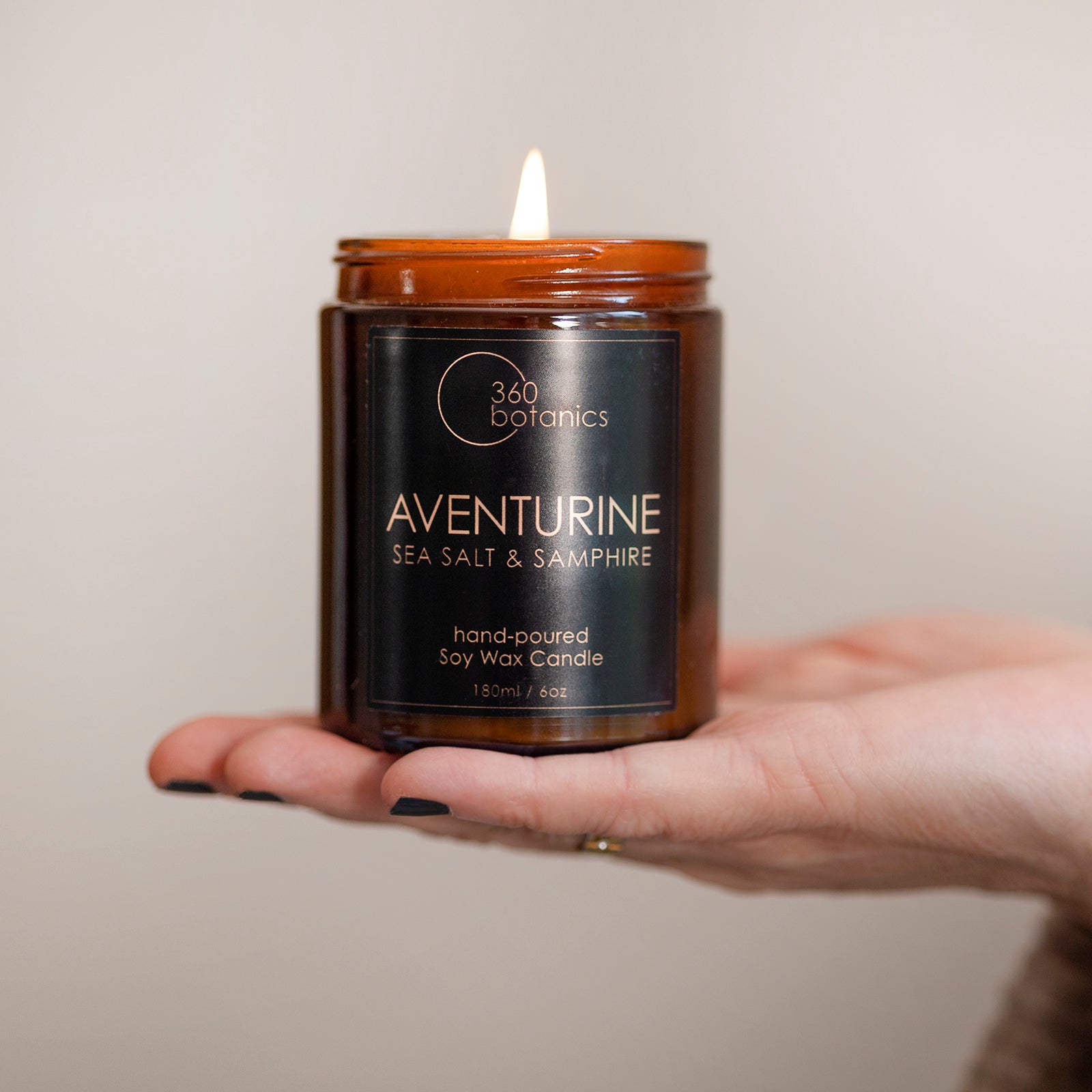 A hand holding a lit Aventurine soy wax candle by 360 Botanics, with a label stating 'Sea Salt & Samphire', 'hand-poured', and a 180ml / 6 oz volume indication, against a neutral background.