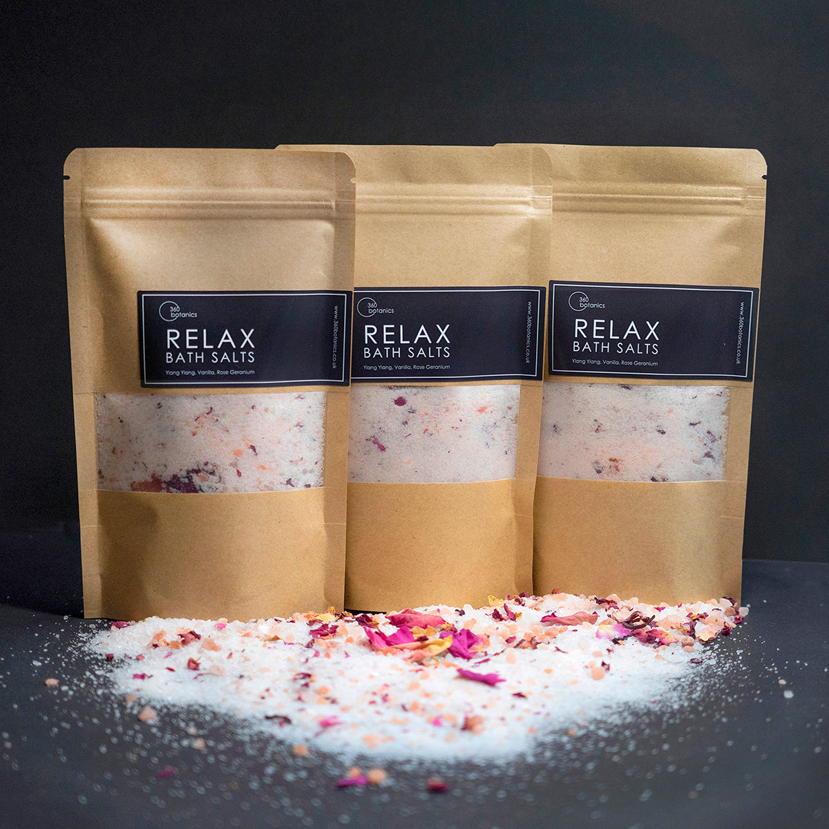 Three kraft paper pouches of '360 Botanics RELAX BATH SALTS' stand in a row against a dark background. The transparent window of each pouch reveals a mix of bath salts infused with petals and herbs. Scattered in front on a black surface is a blend of the colorful salts and botanicals, highlighting the natural ingredients of Ylang Ylang, Vanilla, and Rose Geranium for a calming bath experience