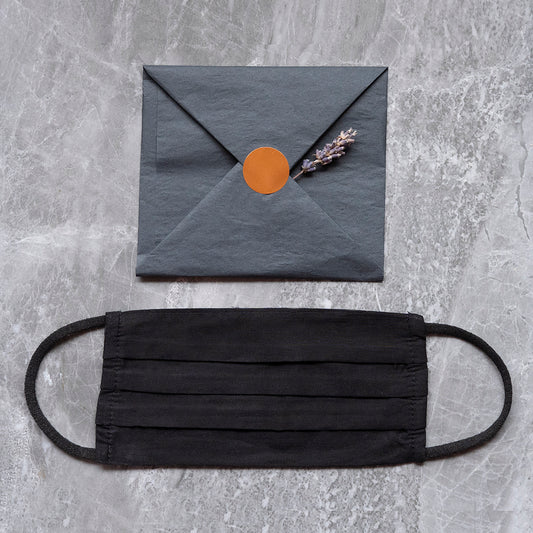  A black fabric face mask laid out on a marble surface below a dark grey envelope sealed with an orange sticker and adorned with a sprig of lavender.
