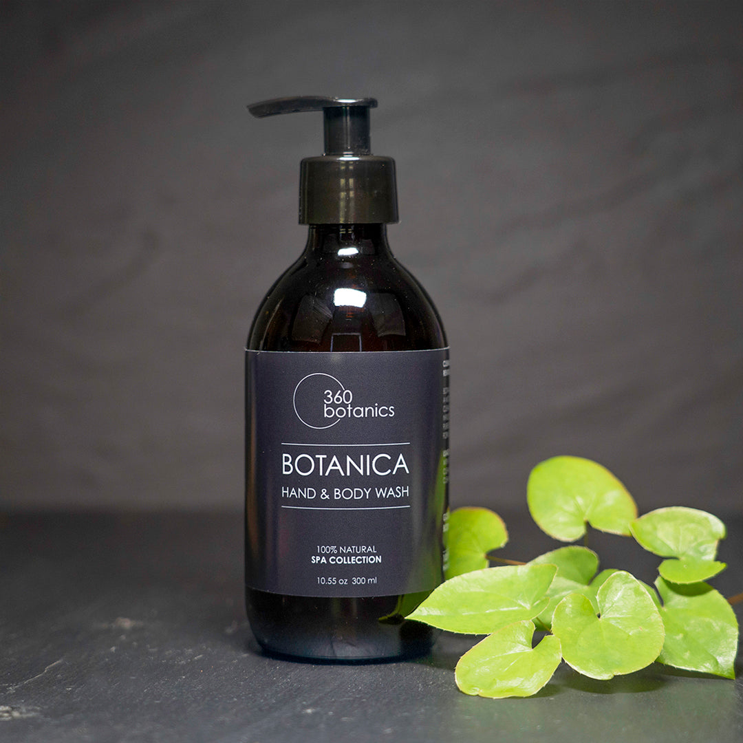  A bottle of 360 Botanics Botanica Hand & Body Wash from the 100% Natural Spa Collection, sized at 10.55 oz or 300 ml, with a pump dispenser. Beside the bottle is a cluster of green leaves set against a dark grey background.