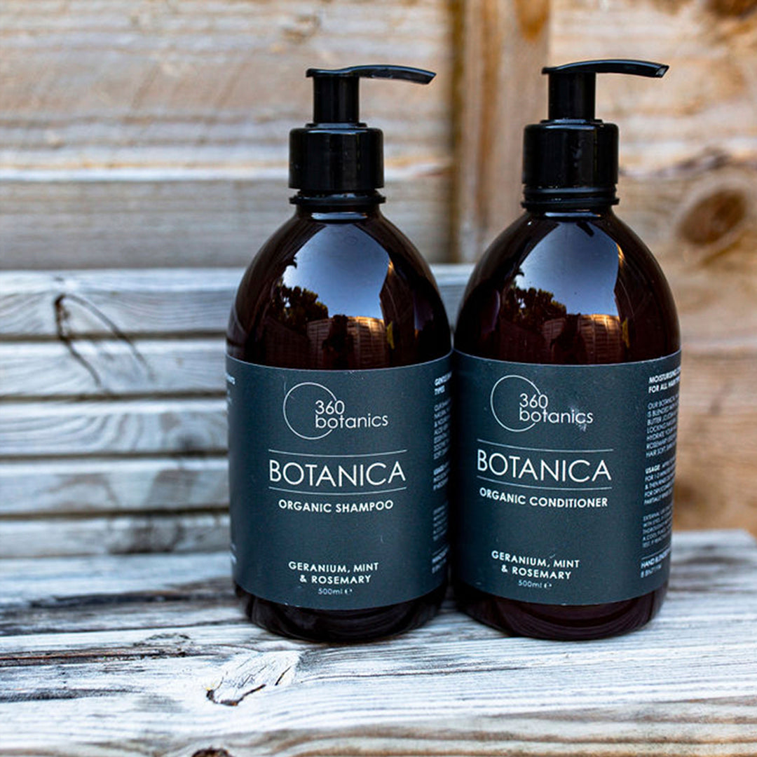 Two 500ml bottles of 360 Botanics Botanica products, one an organic shampoo and the other an organic conditioner, with geranium, mint, and rosemary scents, placed on a rustic wooden surface.