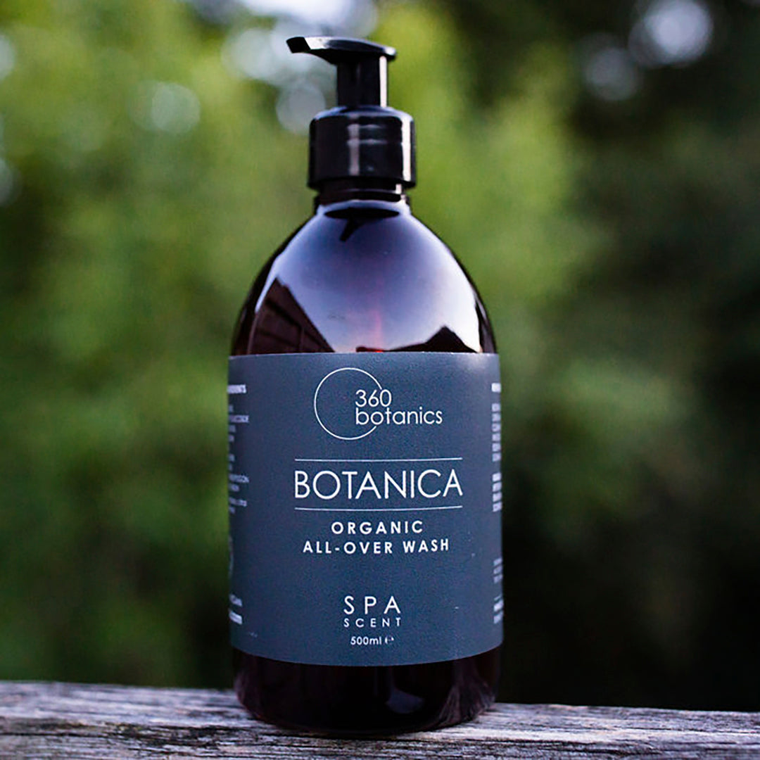 A bottle of 360 Botanics Botanica Organic All-Over Wash with a pump dispenser, featuring "SPA SCENT" and a 500ml volume indication, set upon a wooden surface with a blurred natural green background.