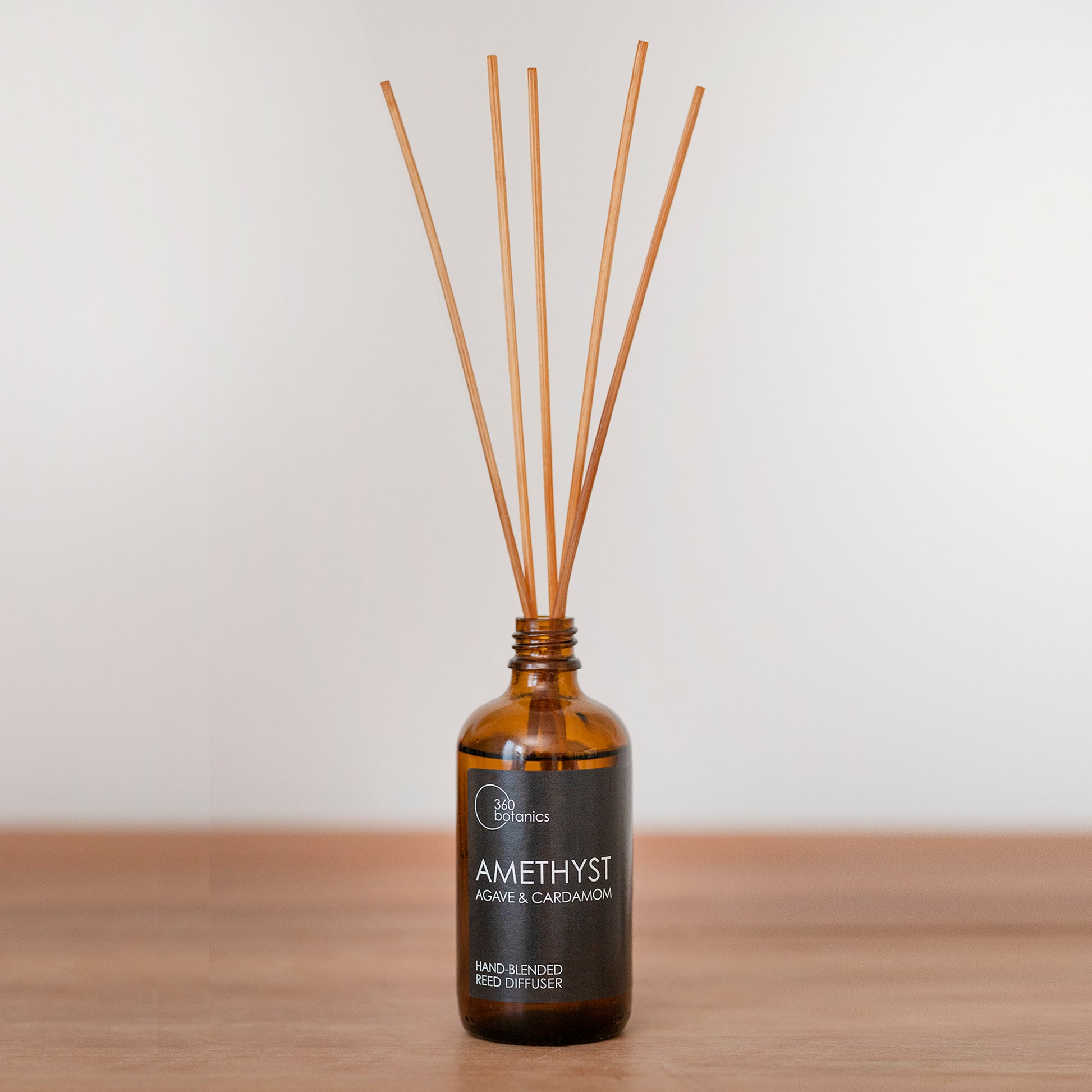 A 360 Botanics Amethyst reed diffuser with agave and cardamom sits on a wooden surface. The amber glass bottle and its array of light brown reeds are set against a soft white background, creating a warm and inviting ambiance, and indicating the product's natural scent profile