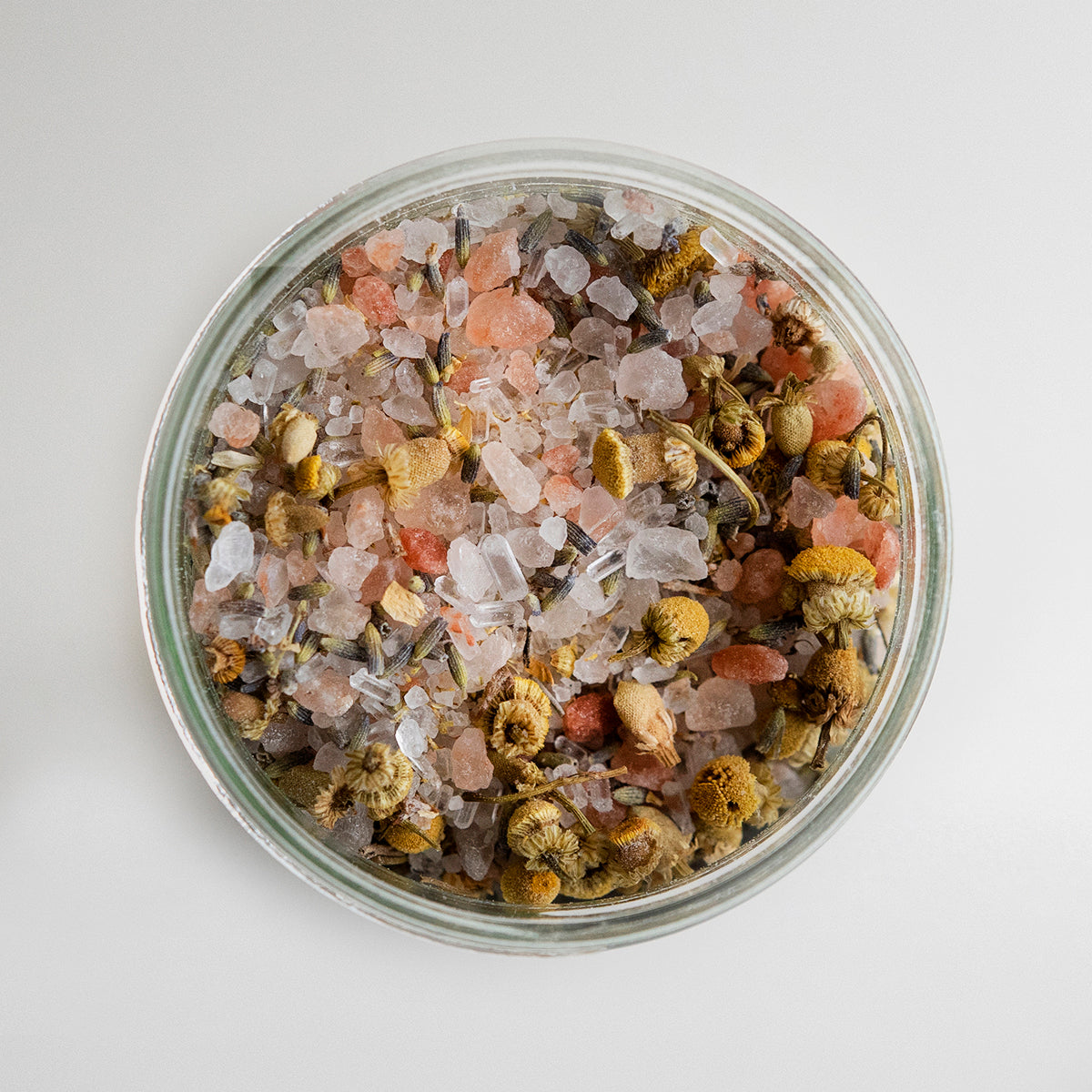 Top view of an open jar filled with a mixture of bath salts and dried herbs, including pink Himalayan salt, clear crystals, and various dried flowers on a white background.
