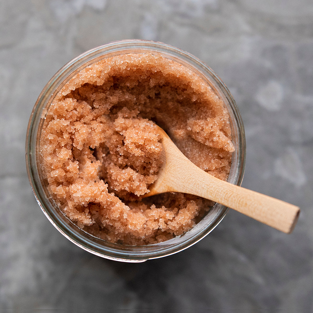 Top-down view of an open jar of 360 Botanics Sugar Scrub, showcasing the granular texture of the natural brown sugar blend. A wooden spoon is nestled in the scrub for application, set against a marble grey background, emphasizing the product's organic and luxurious essence