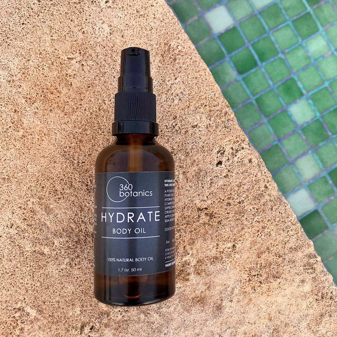 A 360 Botanics HYDRATE Body Oil bottle with a black label, situated on a textured tan ledge with a green tiled pool visible in the blurred background.