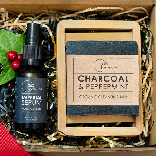 A gift box containing a bottle of 360 Botanics Imperial Serum men's facial oil and a Charcoal & Peppermint organic cleansing bar, both nestled in decorative straw. The box is adorned with holly berries and leaves, suggesting a festive or holiday theme.