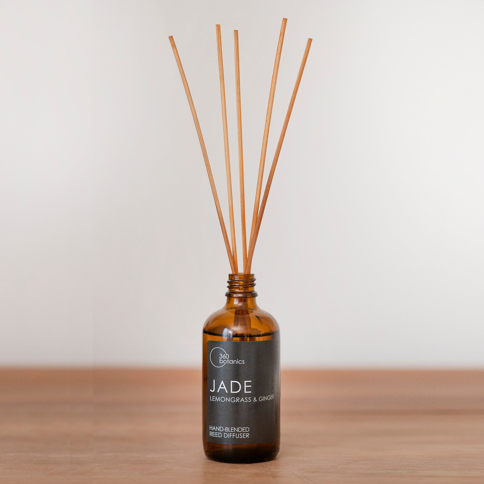360 Botanics Jade reed diffuser with a lemongrass and ginger scent in a brown glass bottle. Several light brown reeds extend upwards, distributing the fragrance. It's presented on a wooden surface against a neutral background, symbolizing the fresh and invigorating essence of the product