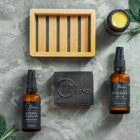 An arrangement of 360 Botanics skincare products on a marble surface, including a bottle of Imperial Serum Men's Facial Oil, a Hydrate Body Oil, and a black soap bar with the embossed logo. A wooden soap dish and a small open jar of product with yellow content add to the natural, organic appeal of the presentation