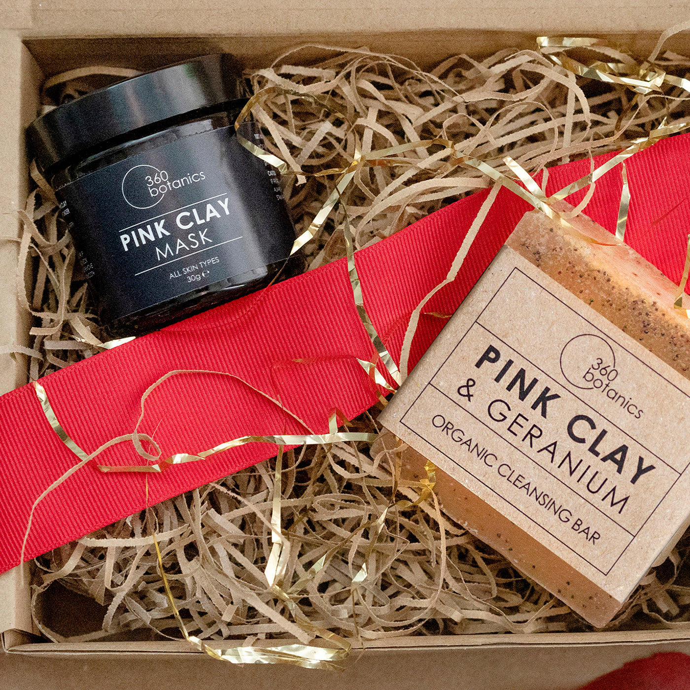 A gift box containing 360 Botanics Pink Clay Mask and Pink Clay & Geranium Organic Cleansing Bar, adorned with red ribbon and natural wood shavings, presenting an elegant skincare set.