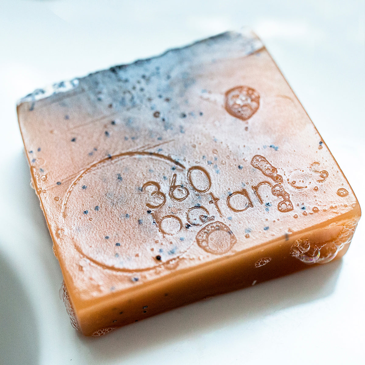 A close-up of a wet 360 Botanics soap bar with the brand logo embossed on its surface, surrounded by delicate bubbles and specks of botanical ingredients. The soap's rich terracotta hue is complemented by a marbled blue top, creating a luxurious and organic aesthetic