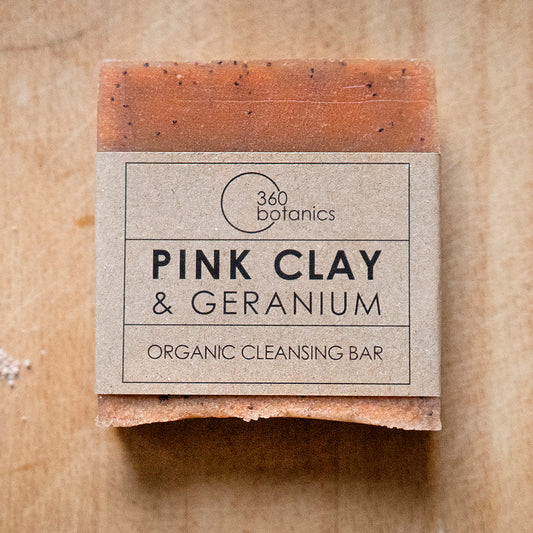360 Botanics Pink Clay & Geranium Organic Cleansing Bar, displayed on a natural wood surface. The soap is a soft terracotta colour with visible flecks of pink clay, wrapped in a kraft paper label that adds to its artisanal and eco-friendly appeal