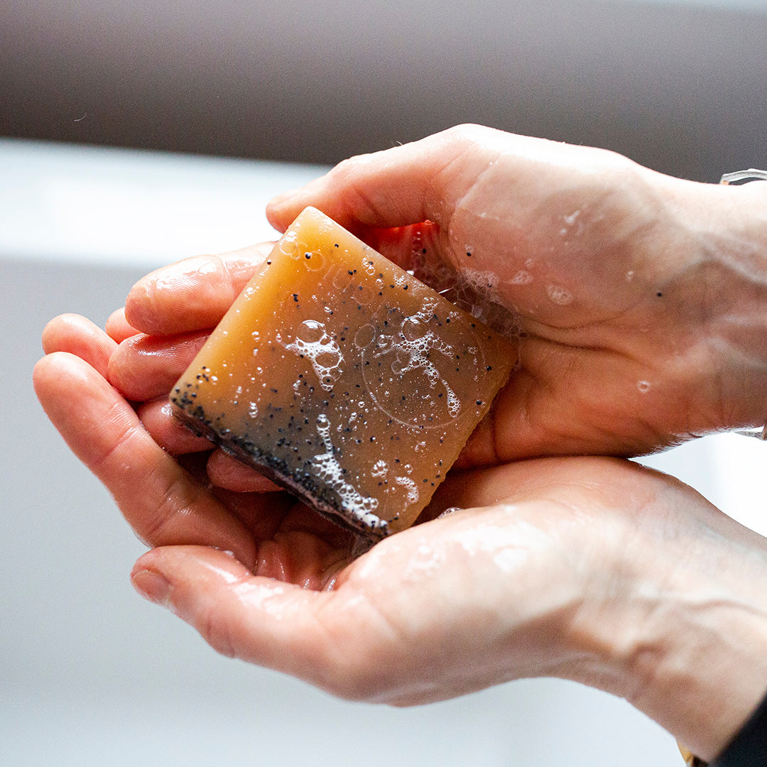 Hands lathering up a square piece of amber-coloured pink clay soap with black scrub particles, against a soft-focus grey background, emphasizing the natural and exfoliating properties of the product