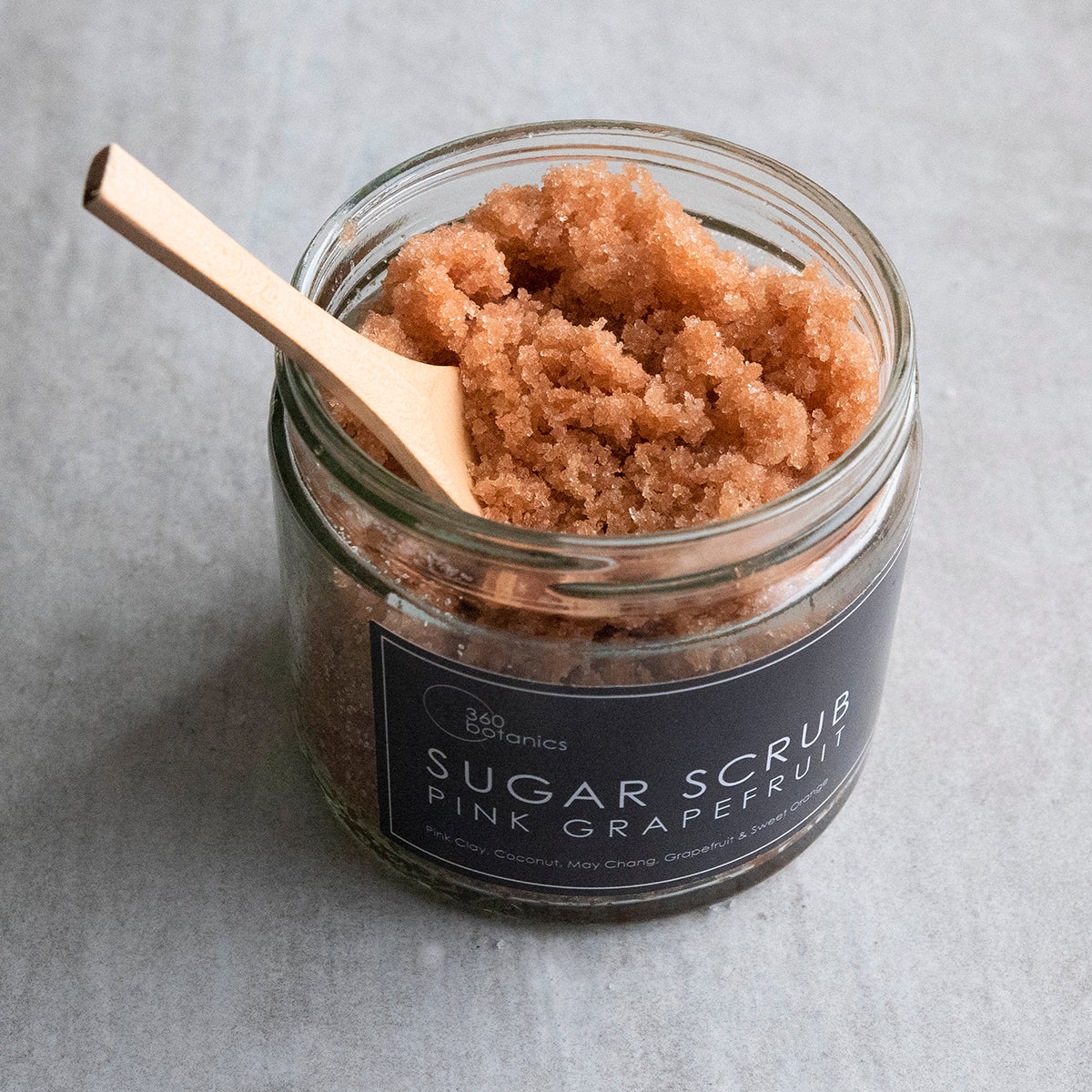 An open jar of 360 Botanics Sugar Scrub with Pink Grapefruit, revealing a rich texture of brown sugar crystals. A wooden spoon rests on top, partially covered by the scrub, set against a grey background, highlighting the product's natural exfoliating qualities