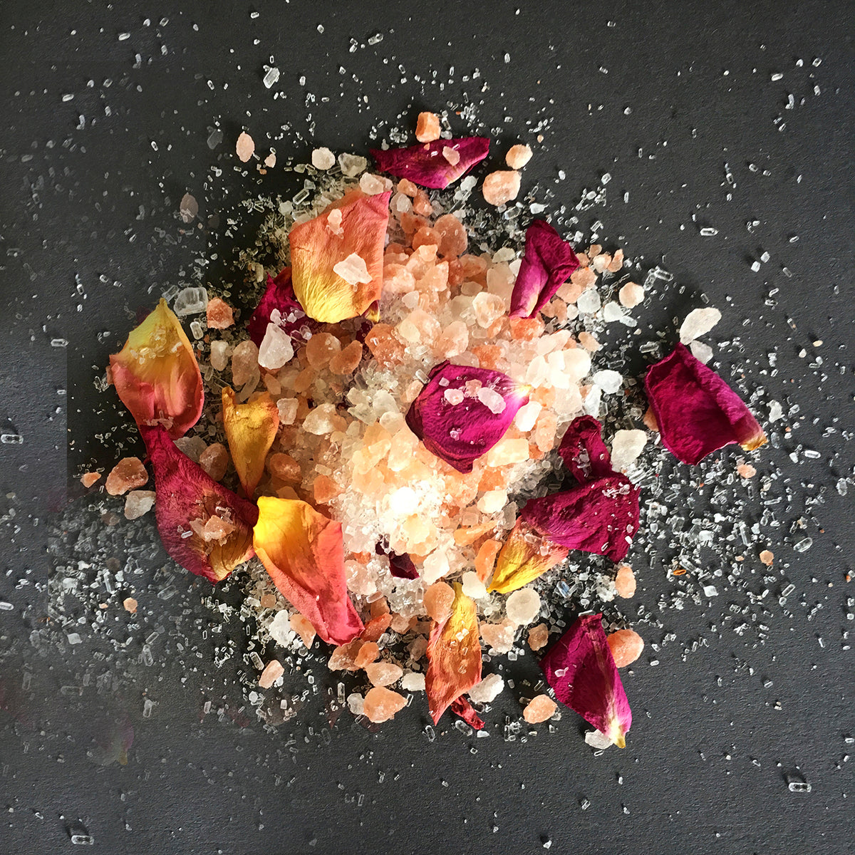  A close-up view of a luxurious mixture of bath salts and dried flower petals arranged on a dark, textured surface. The salt crystals vary in color from clear to peach and amber tones, while the flower petals are rich in color, including shades of deep burgundy, bright yellow, and warm orange. The scattered arrangement gives an impression of a relaxing and aromatic bath experience