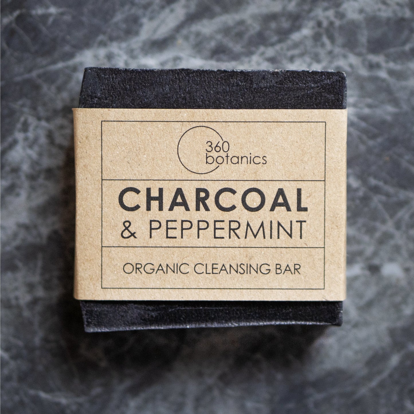 Close-up view of a 360 Botanics organic cleansing bar packaging, featuring 'CHARCOAL & PEPPERMINT' on a natural kraft paper label. The black bar of soap is partially visible, suggesting the product's natural charcoal ingredient, all set against a grey marbled background, evoking a clean and organic aesthetic