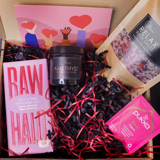 The image shows a curated gift box containing various wellness items. Inside, there is a "360 botanics" AMETHYST 100% vegan soy candle in a black jar, a bar of "RAW HALO" Mylk & Pink Himalayan Salt organic chocolate, a packet of "PUKKA" Love Organic tea, and a packet of "Relax" bath salts from 360 Botanics. The box is filled with black and red shredded paper, and there's a greeting card with a design of hands holding hearts at the top. The composition suggests a thoughtful, health-conscious gift set.