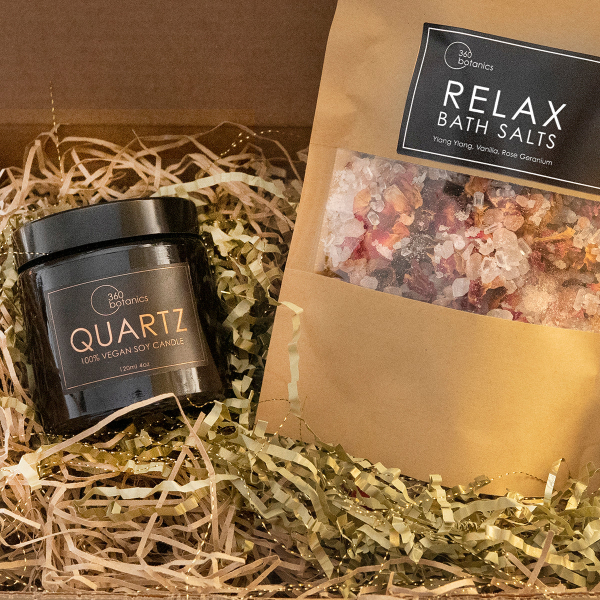 A cosy and inviting gift set from 360 Botanics, presented in a brown cardboard box filled with natural shredded paper. Inside, there's a sleek black glass jar labeled 'QUARTZ 100% VEGAN SOY CANDLE' and a kraft paper pouch of 'RELAX BATH SALTS' with visible crystals of Ylang Ylang, Vanilla, and Rose Geranium, promising a soothing and aromatic experience