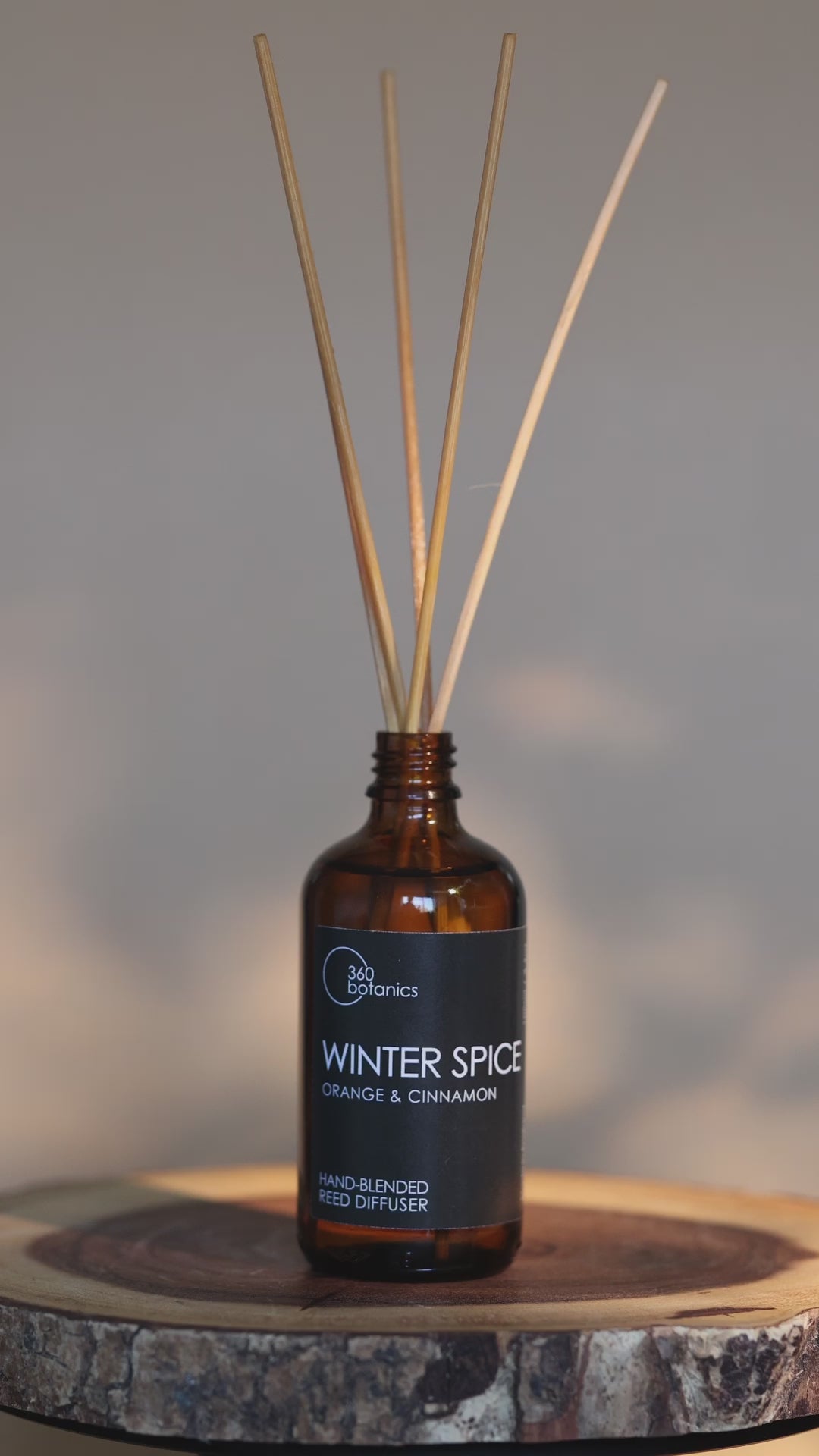 An atmospheric video of the 360 Botanics Winter Spice reed diffuser with orange and cinnamon, placed on a wooden coaster. The amber bottle with light brown reeds is highlighted by a soft, warm light in the background, invoking a cozy winter ambiance