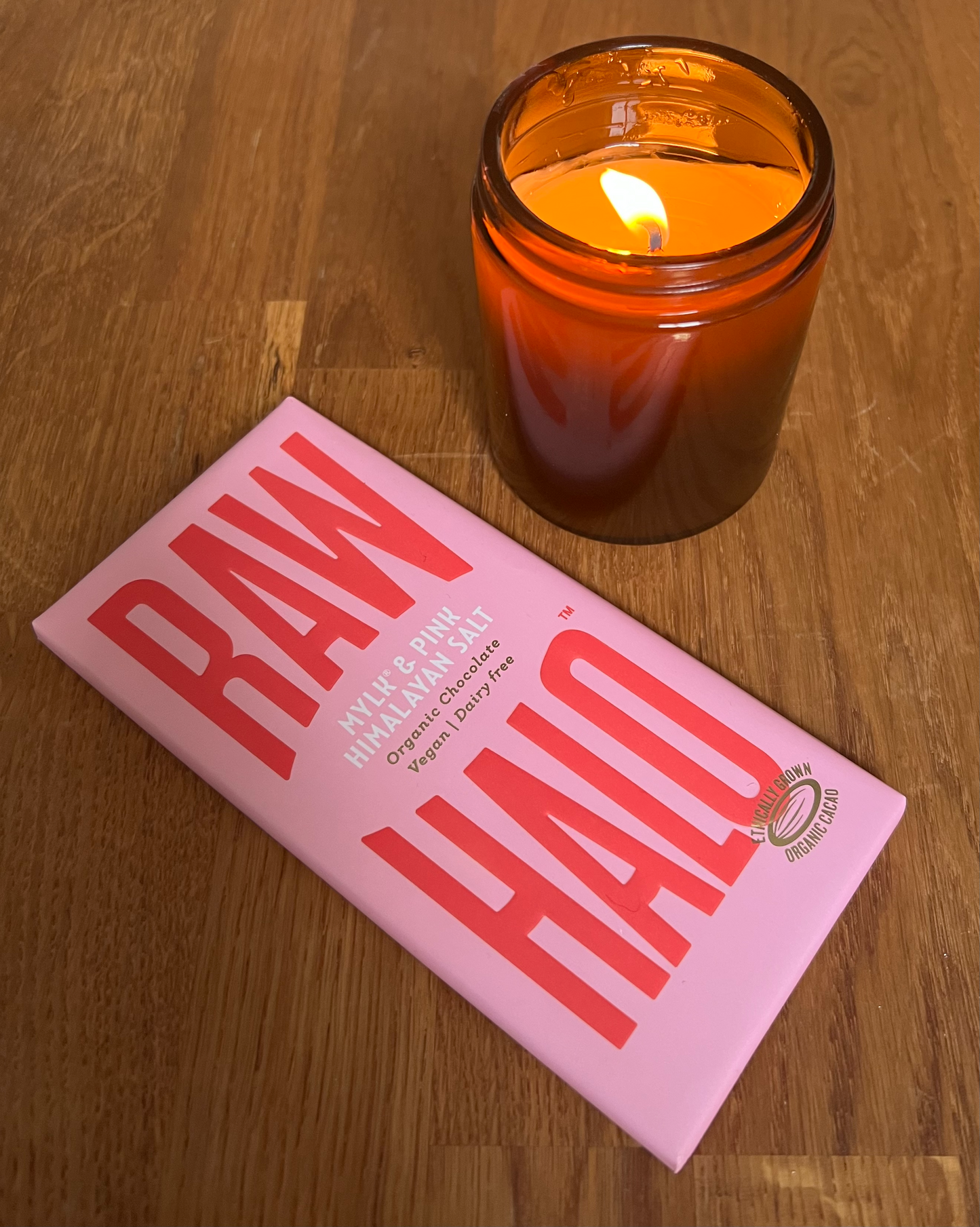  The image depicts a lit amber glass candle next to a pink RAW HALO Mylk & Pink Himalayan Salt organic chocolate bar. The candle is on the right, glowing warmly on a wooden surface, while the chocolate bar lies flat on the left with prominent red branding. The scene suggests a cozy and indulgent atmosphere.