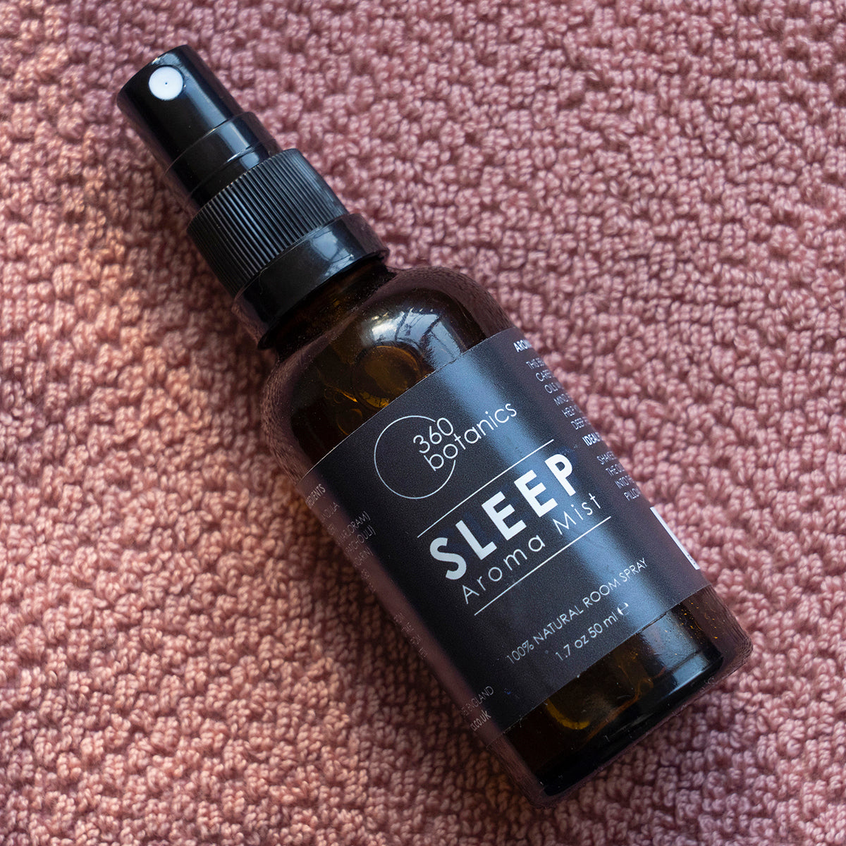 A bottle of 360 Botanics Sleep Aroma Mist, a 100% natural room spray in a 1.7 oz or 50 ml bottle with a spray nozzle, resting on a textured pink fabric surface.