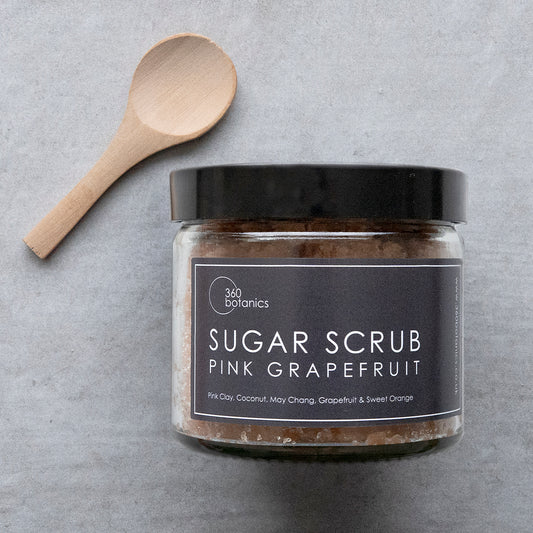 A clear jar labeled '360 Botanics SUGAR SCRUB PINK GRAPEFRUIT' with ingredients like Pink Clay, Coconut, May Chang, Grapefruit, and Sweet Orange listed. Beside the jar is a wooden spoon, all set against a textured grey background, showcasing the product's natural ingredients and hinting at a refreshing citrus aroma