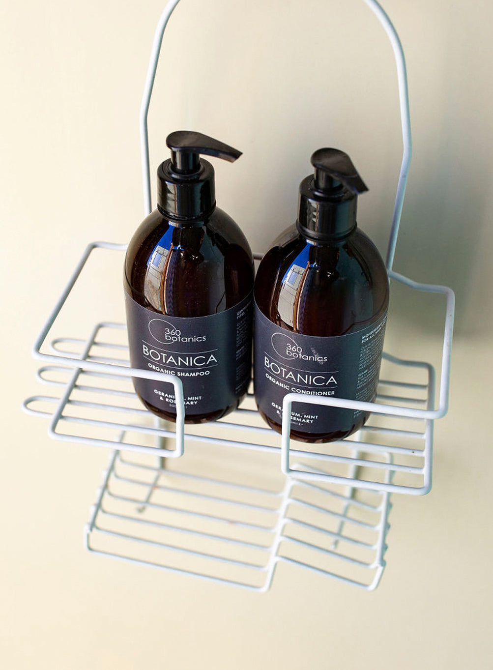 Two bottles of 360 Botanics Botanica products on a wire shelf: one is an organic shampoo and the other an organic conditioner, both featuring eucalyptus, mint, and rosemary, placed against a light-colored wall.