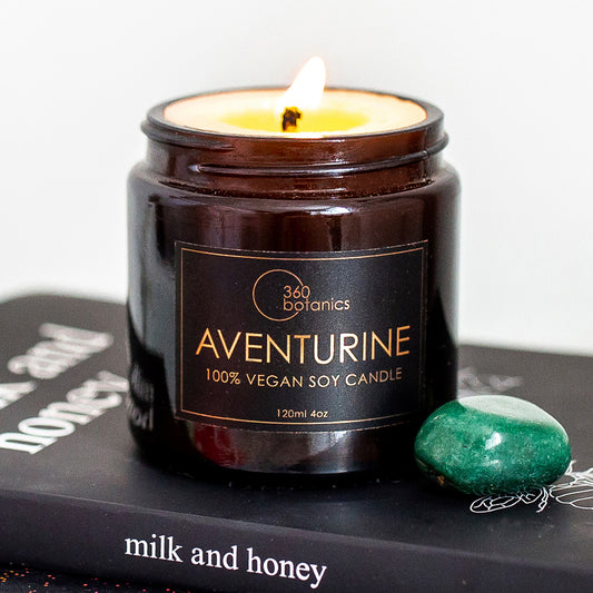  A lit 100% vegan soy candle by 360 Botanics named Aventurine, with a visible flame, placed on a book titled 'milk and honey', next to a polished green aventurine gemstone