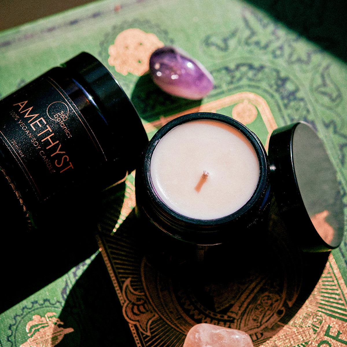  An Amethyst crystal-inspired soy wax candle from 360 Botanics, with a single wick, displayed next to a purple crystal on a green decorative surface.